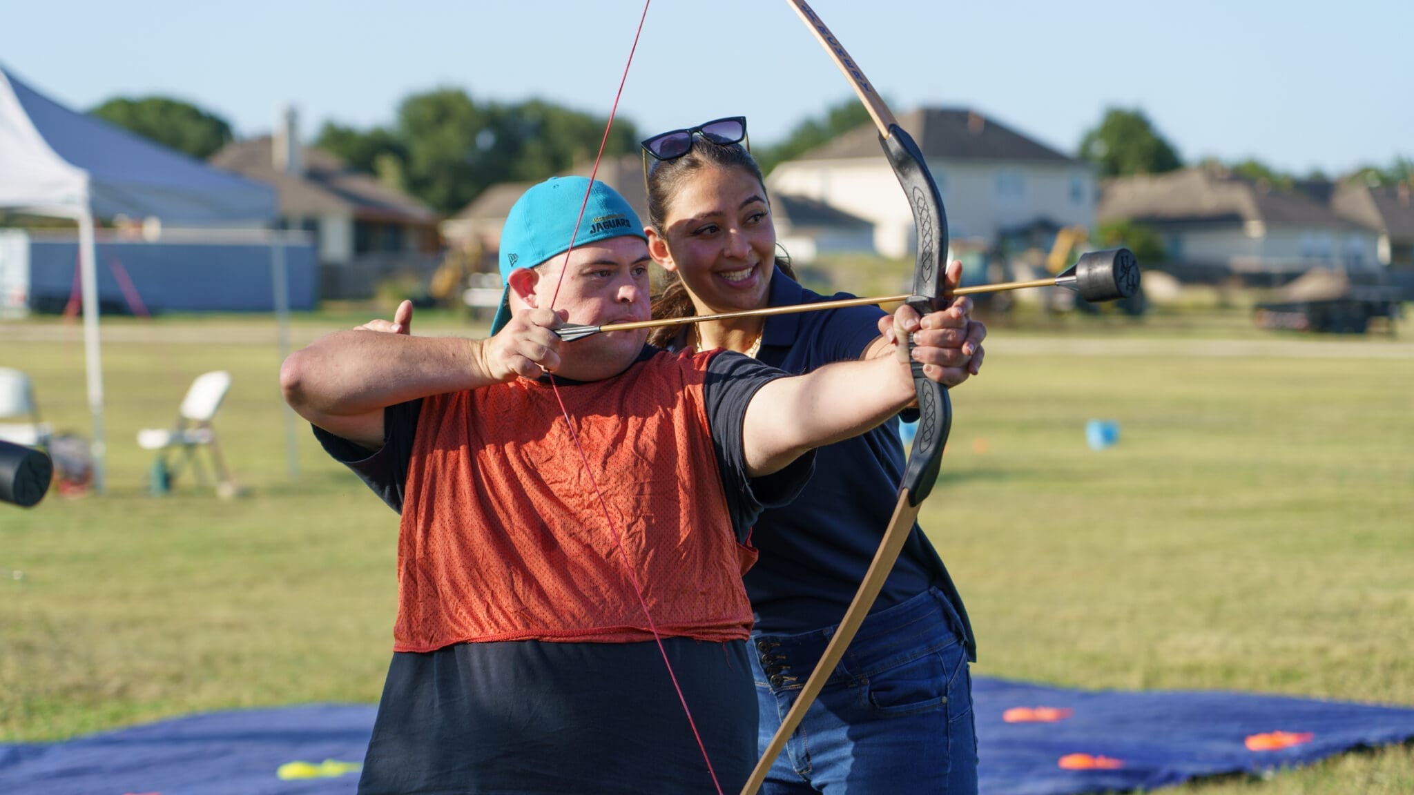 A young man wearing an orange vest and blue cap aims a bow and arrow with the help of a smiling woman standing behind him on a grassy field. Houses and outdoor event equipment are visible in the background.