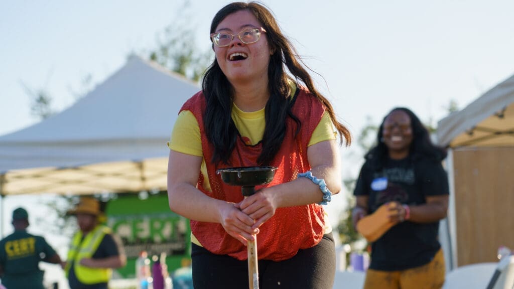 A young woman with Down syndrome smiles and participates in an outdoor event, balancing a black object on a wooden stick. People and white tents are visible in the background. The atmosphere appears joyful and energetic.