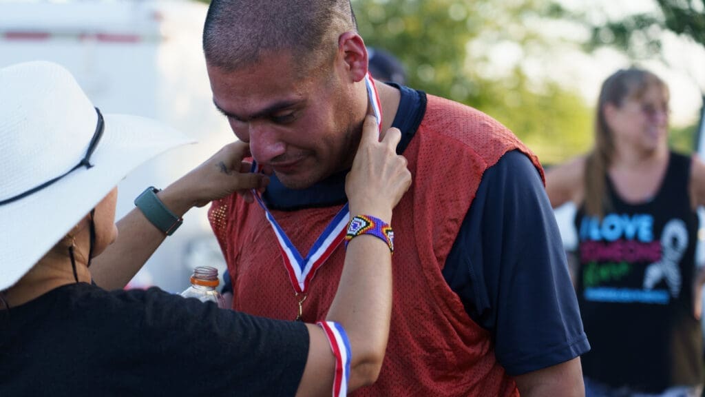 A person places a medal around a man's neck, who appears emotional. He’s wearing a red sports bib, and the scene is outdoors with others in the background, including a person in a “Love” shirt.
