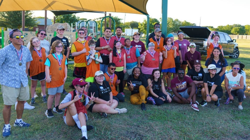A large group of people, some wearing orange jerseys and medals, pose and smile together outdoors under a canopy on a grassy field. Playground equipment and a parked car are visible in the background.