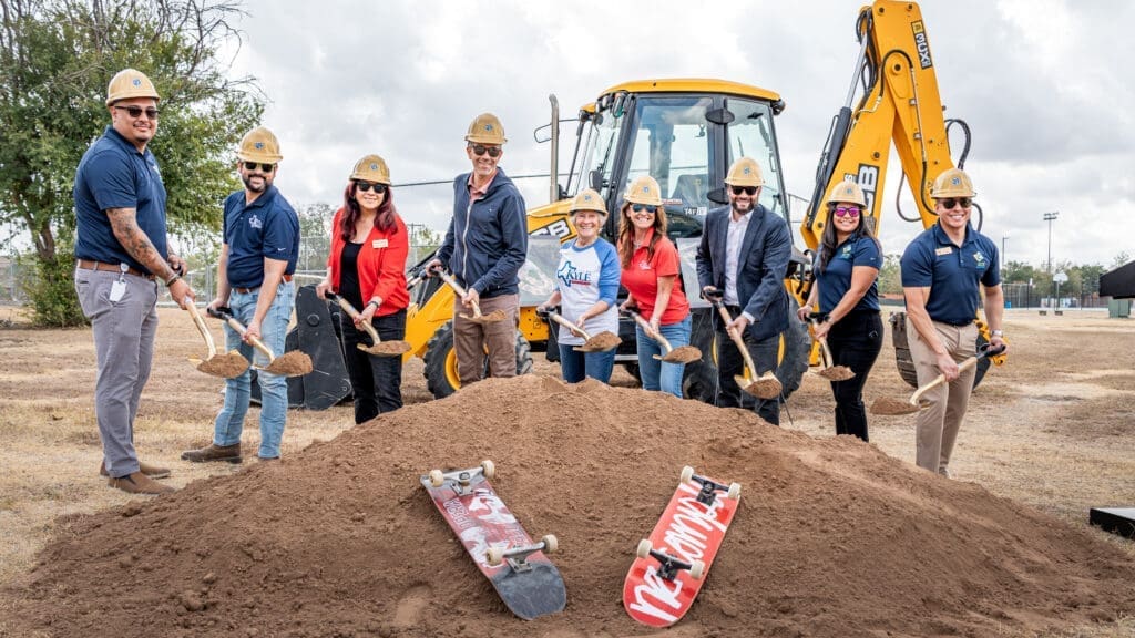 A group of people in hard hats and safety gear stand in front of a backhoe, smiling and holding shovels over a mound of dirt with three skateboards placed on it, during a groundbreaking ceremony outdoors.