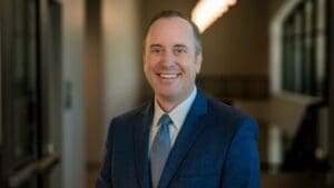 City Manager Bryan Langley in a blue suit and tie smiles at the camera in a hallway with soft lighting and blurred background.