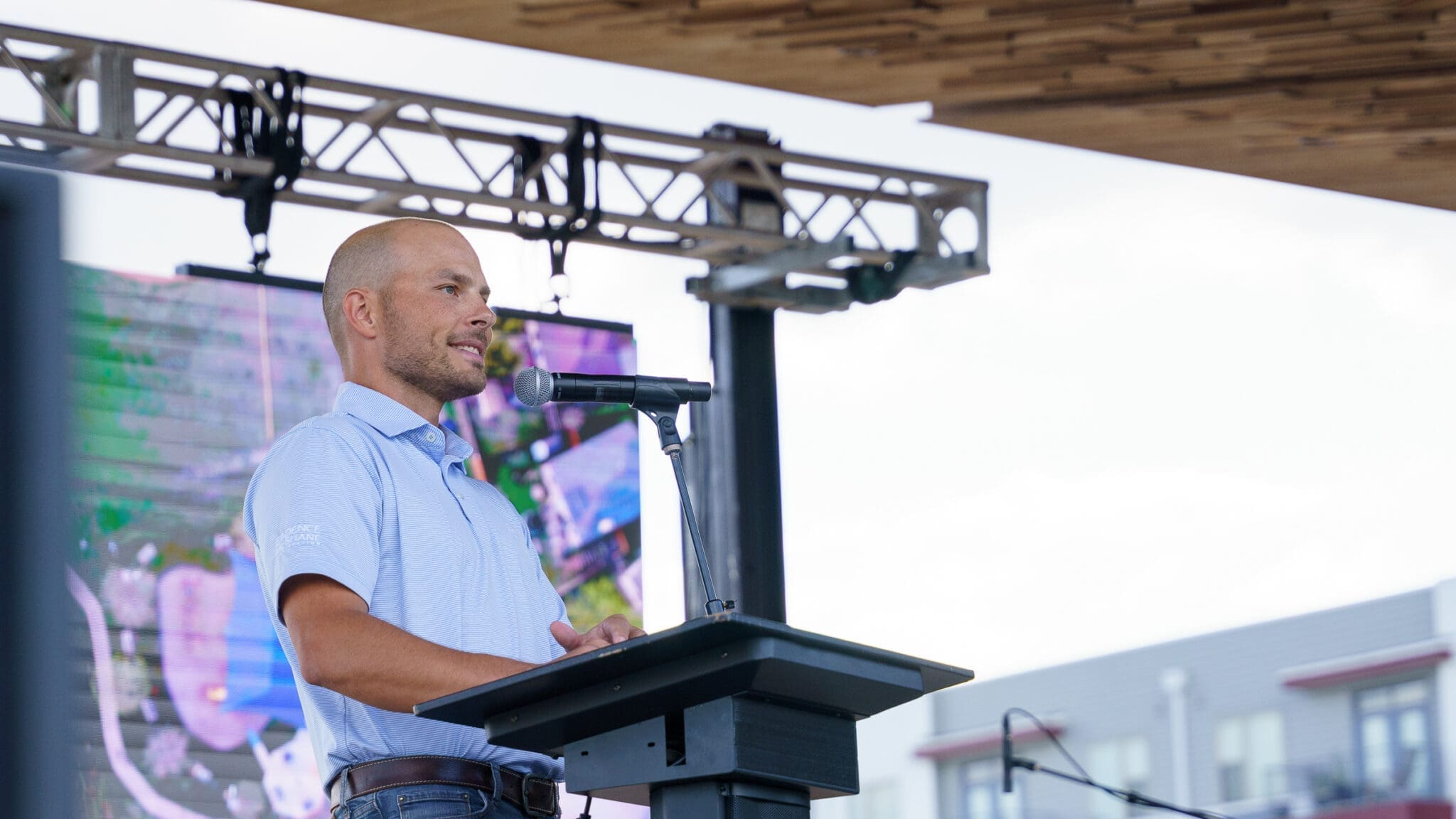 A man in a light blue shirt speaks at a podium with a microphone on an outdoor stage. There is a large screen and buildings in the background.