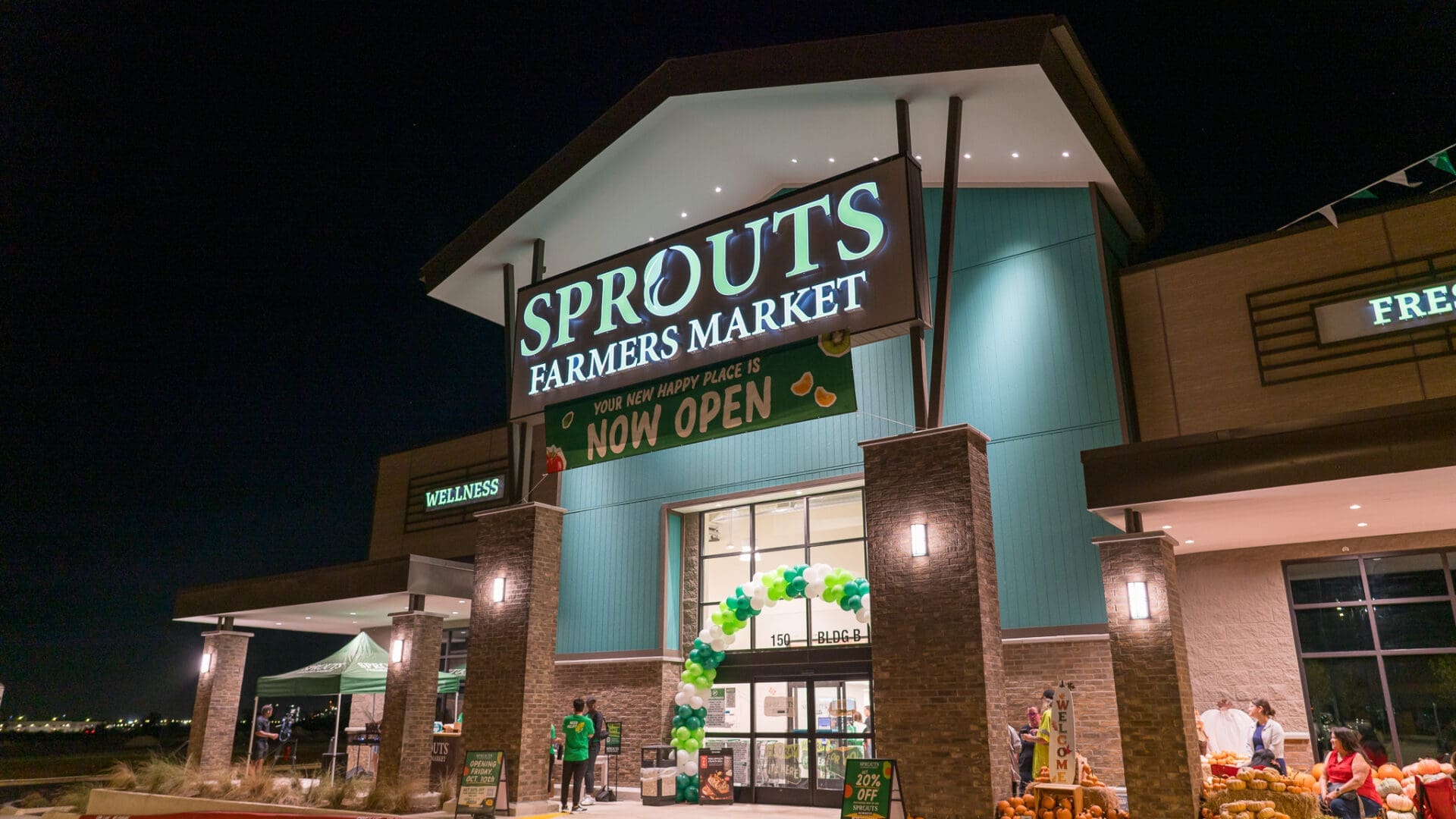 The exterior of a brightly lit Sprouts Farmers Market store at night, with a “Now Open” sign and green and white balloon arch at the entrance.
