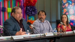 Three people sit at a table with microphones, speaking at an event decorated with colorful paper flowers and banners. The man on the left is gesturing while speaking; the other two listen attentively.