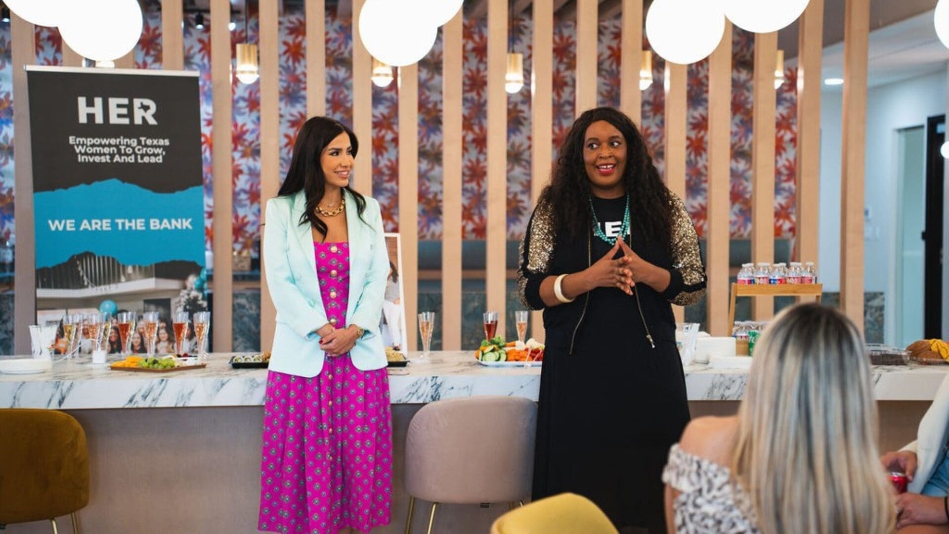 Two women stand in front of a table with food, speaking to a small audience at an indoor networking event. A banner reading "HER Empowering Texas Women to Grow, Invest And Lead" is displayed beside them.