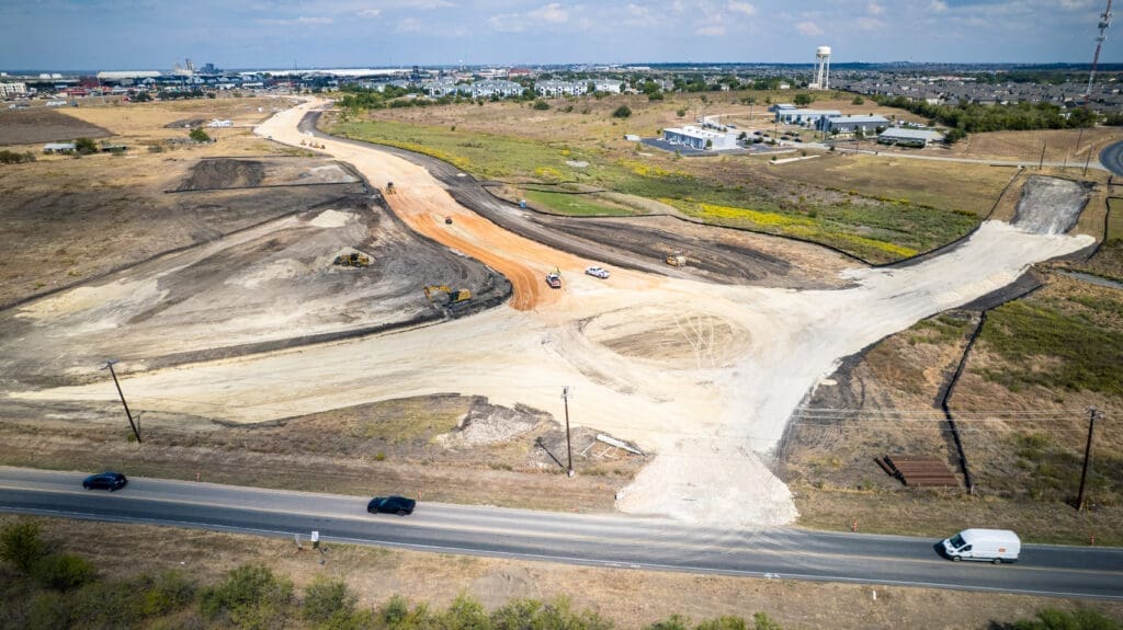 Aerial view of a road under construction in a dry, open area. Construction vehicles are working on the site, with unfinished roads and cleared land visible. Nearby, buildings and roads are surrounded by open fields.