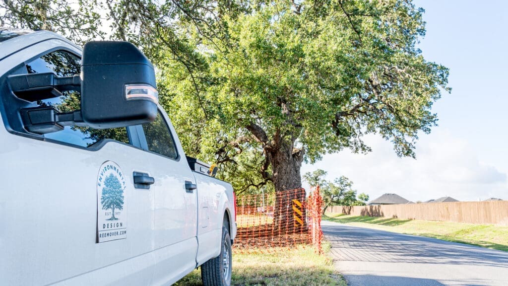 A white pickup truck with a tree care company logo is parked beside a large leafy tree, next to an orange construction fence by a suburban road under a sunny sky.