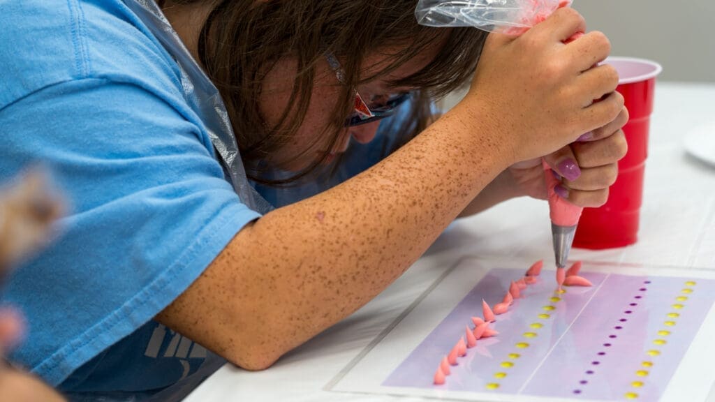 A person with long brown hair uses a piping bag filled with pink icing to make decorative shapes on a practice sheet with colorful dots, focusing intently on their work.