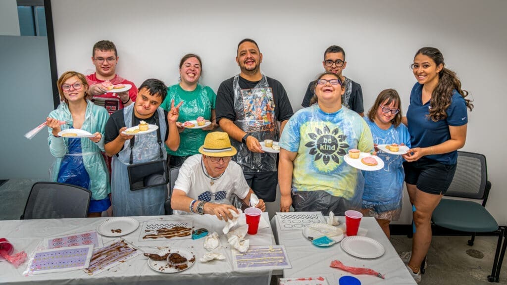 A group of smiling people of various ages, some wearing aprons, stand and sit around a table decorated with art supplies and snacks. They proudly display decorated cookies and appear to be enjoying a fun, creative activity together.