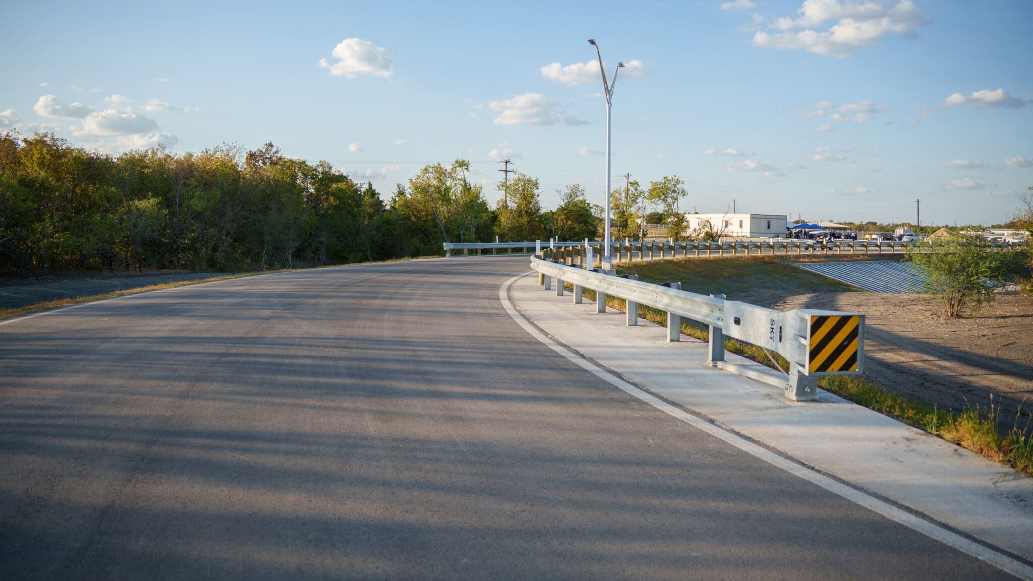 A curved, paved road bordered by metal guardrails runs through a natural area with trees on the left. The sky is mostly clear with scattered clouds, and a few buildings are visible in the background.