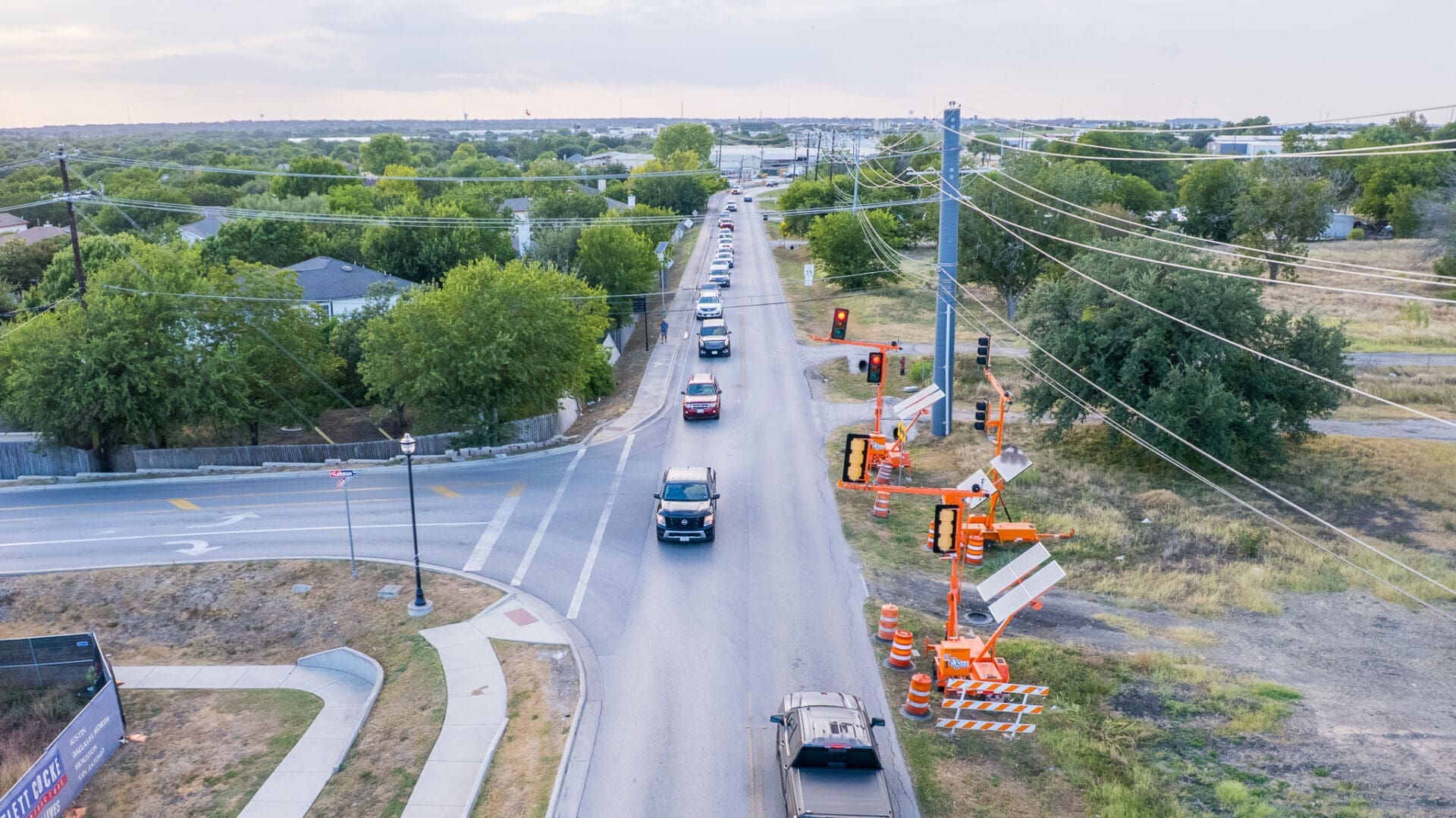 Aerial view of cars lined up in traffic on a two-lane road, with construction signs, orange cones, and traffic lights on one side. Trees and houses are visible on both sides of the road.