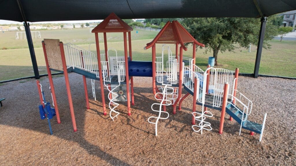 A playground with slides, climbing structures, and ladders under a shade canopy. The ground is covered with wood chips, and there is grass and trees in the background.