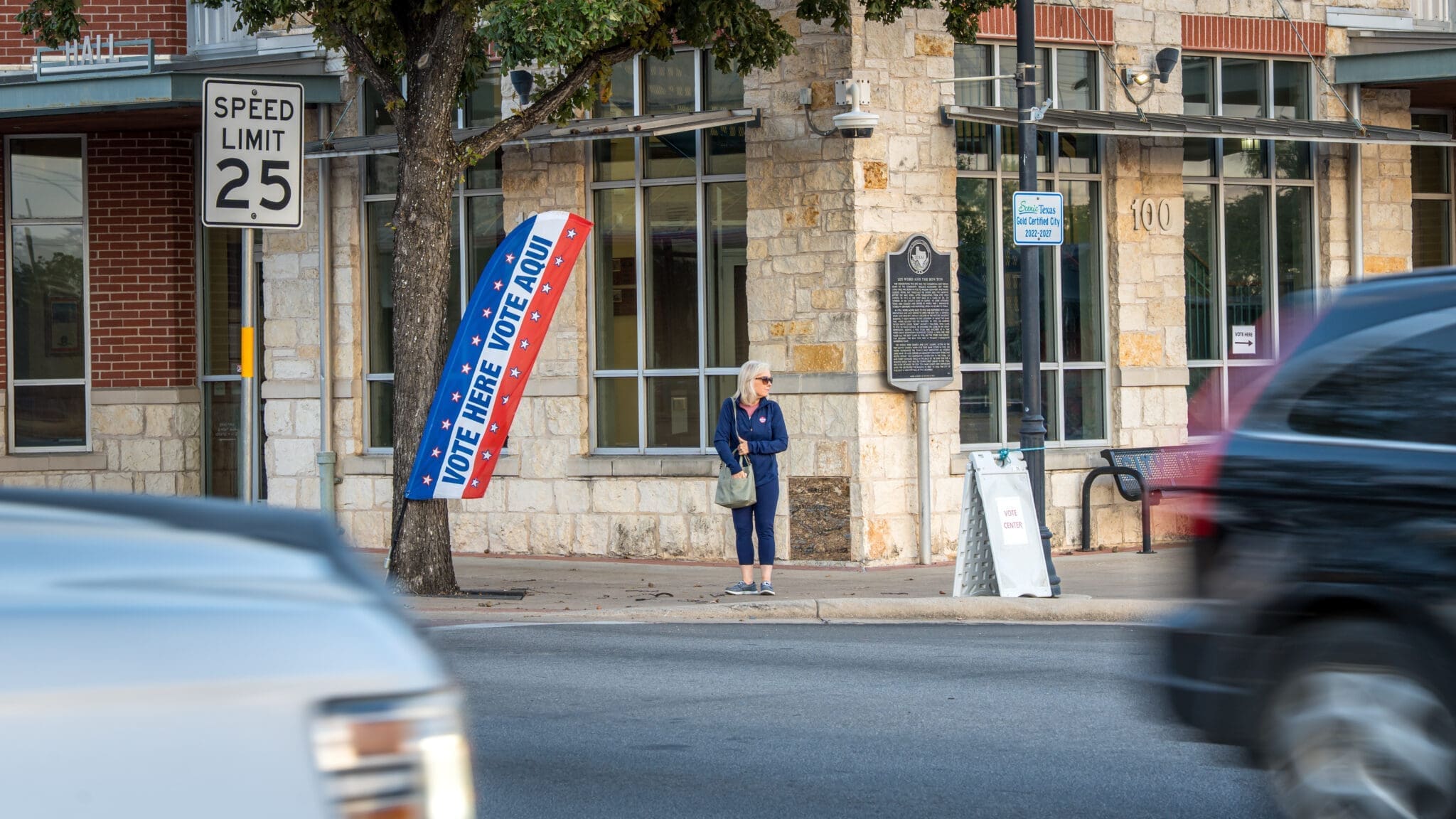 A woman stands on a sidewalk near a polling place sign reading "VOTE HERE - VOTE AQUÍ," with cars passing by on the street and a speed limit 25 sign visible.