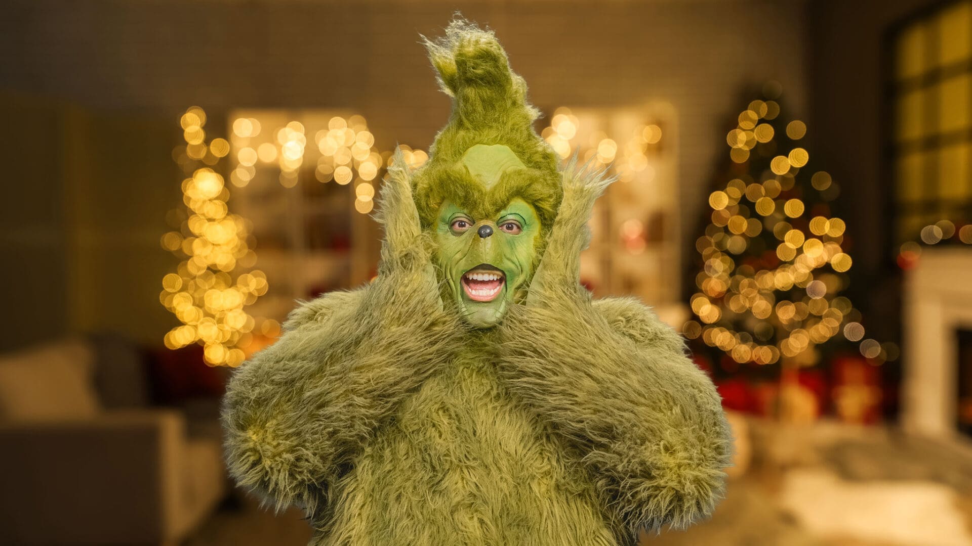A person dressed as the Grinch in a furry green costume stands indoors with hands on their face; the background features blurry, festive Christmas lights and decorated trees.