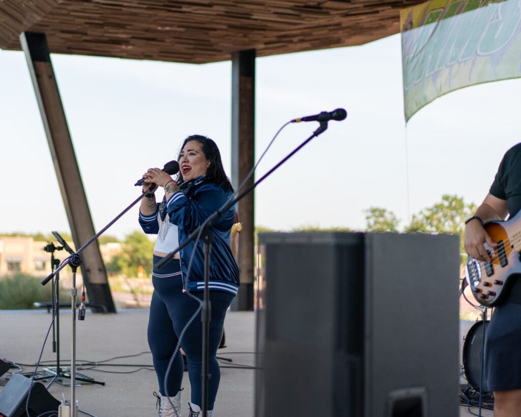 A woman in a blue jacket sings into a microphone on an outdoor stage, with a musician playing bass guitar beside her. Speakers and microphones are visible, and a green banner hangs in the background.