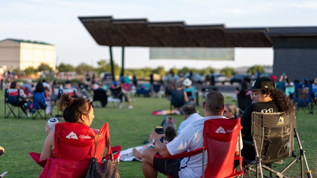 People sit on lawn chairs at an outdoor event, enjoying a sunny day. A stage is visible in the background and groups are scattered across the grass, suggesting a relaxed, communal atmosphere.