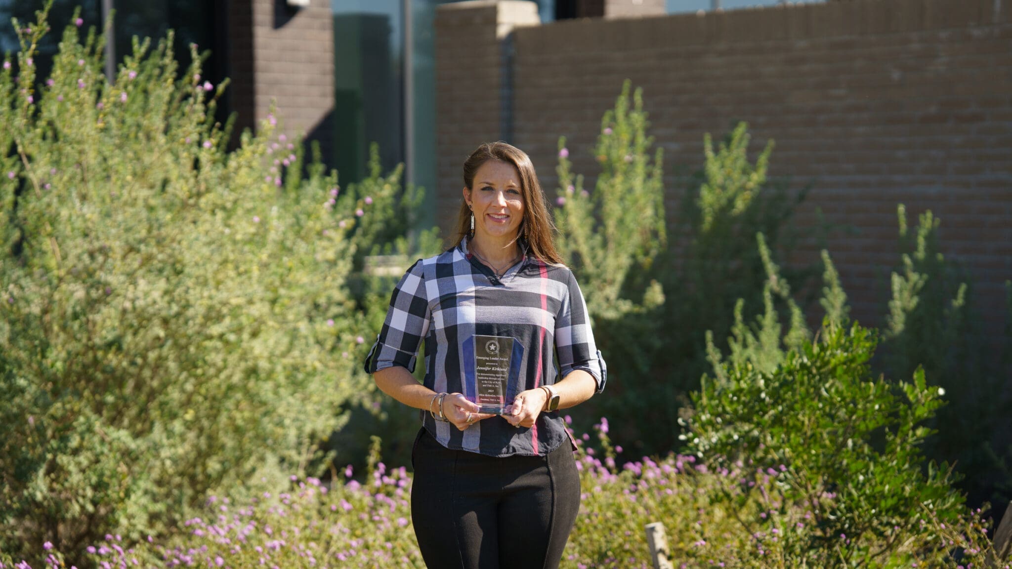 A woman stands outdoors in front of green shrubs, holding a glass award and smiling at the camera. She is wearing a plaid shirt and black pants. Sunlight shines on the scene.