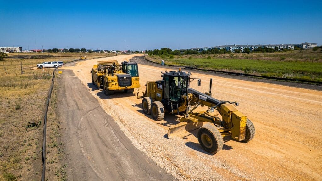 A construction site with large yellow machinery, including a grader and a dump truck, working on leveling a wide gravel road; grassy fields and buildings are visible in the background under a clear blue sky.