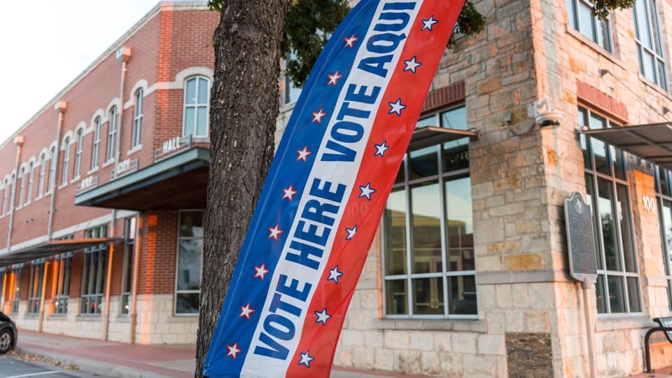 A red, white, and blue feather flag with stars displays the words "VOTE HERE VOTE AQUI" in front of a brick and stone building on a sunny day.