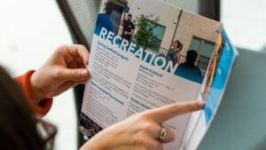 A person holds and points to a brochure listing recreational activities, including spring softball leagues, adult kickball, and tennis lessons, with a large "RECREATION" heading at the top.