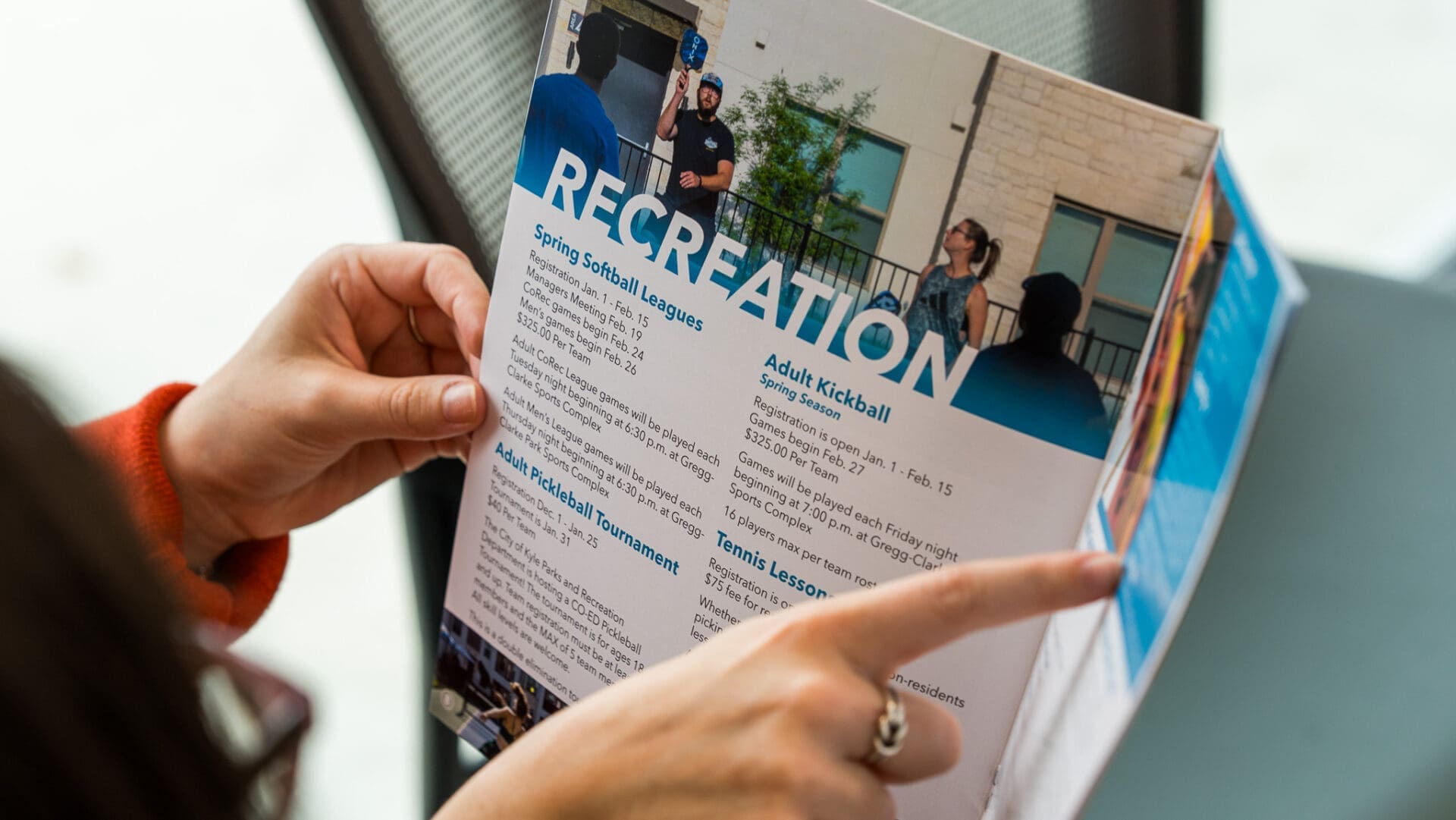 A person holds and points to a brochure listing recreational activities, including spring softball leagues, adult kickball, and tennis lessons, with a large "RECREATION" heading at the top.