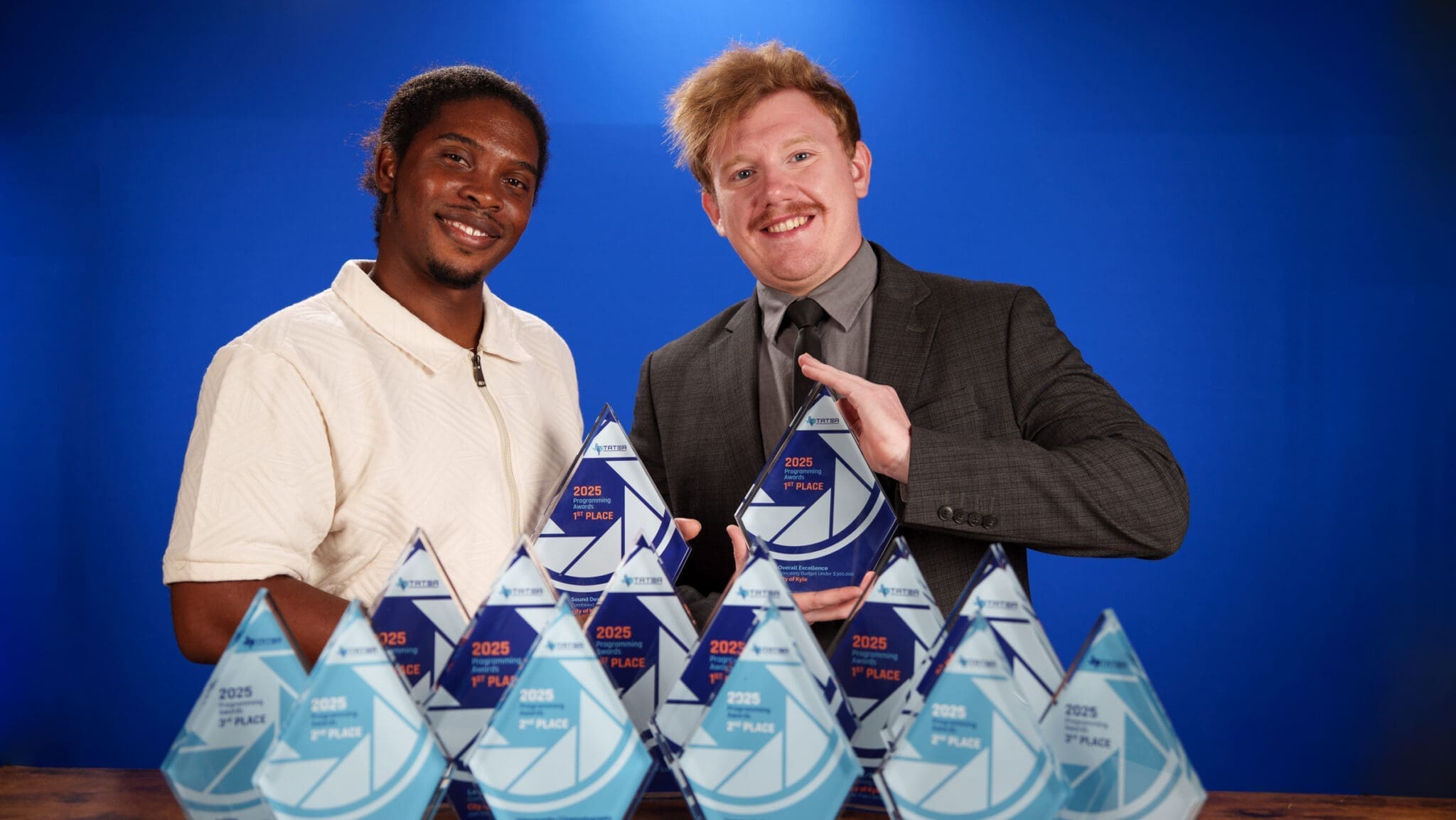 Two smiling men pose in front of a blue background, each holding a blue triangular award. Several similar awards are arranged on the table in front of them. One man wears a light shirt, and the other wears a suit.