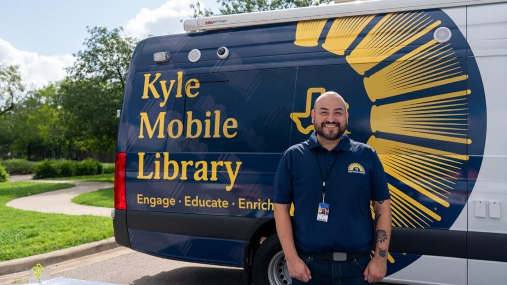 A smiling man stands in front of the Kyle Mobile Library van, which displays the words “Engage • Educate • Enrich” on its side. Trees and a sunny sky are visible in the background.