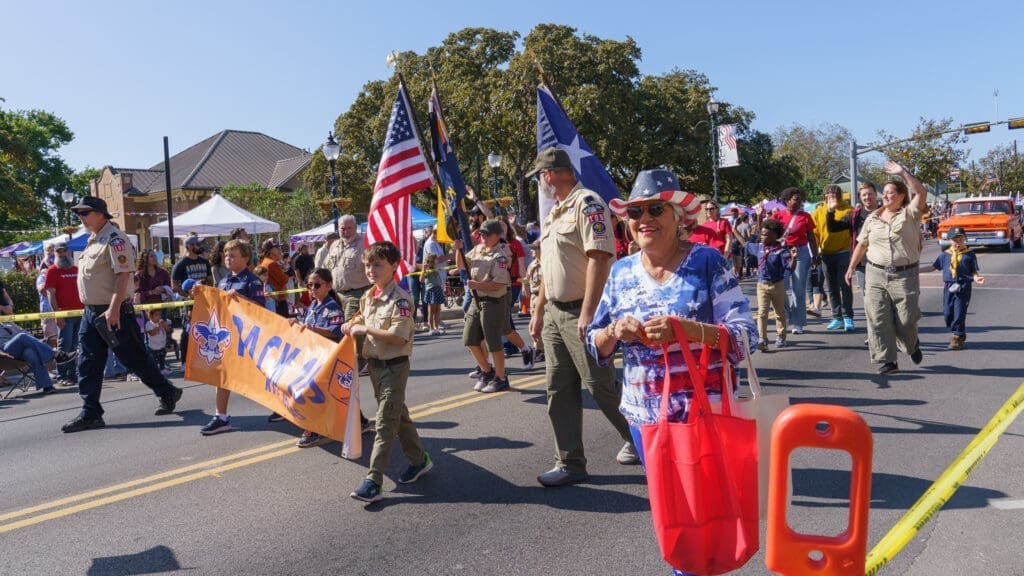 A group of scouts and adults holding flags and a banner march in a sunny parade. Spectators watch from the sidelines. One woman in front wears a hat and sunglasses, carrying a red bag, and smiles at the camera.