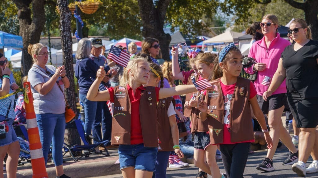 A group of girls in brown vests march in a parade, waving small American flags. Adults and onlookers stand nearby, clapping and watching. The event takes place outdoors under sunny skies.