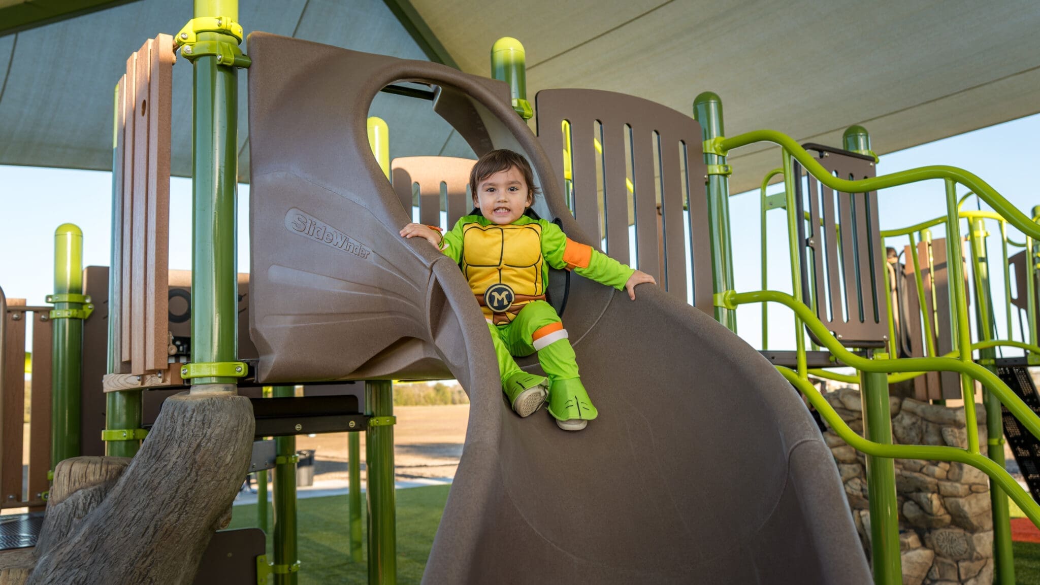 A young child wearing a Teenage Mutant Ninja Turtles costume slides down a tan plastic slide at a playground with green and brown play structures under a large canopy.