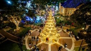 A large Christmas tree adorned with golden lights stands in the center of a lively plaza at night, surrounded by people and festive buildings decorated with bright holiday lights.