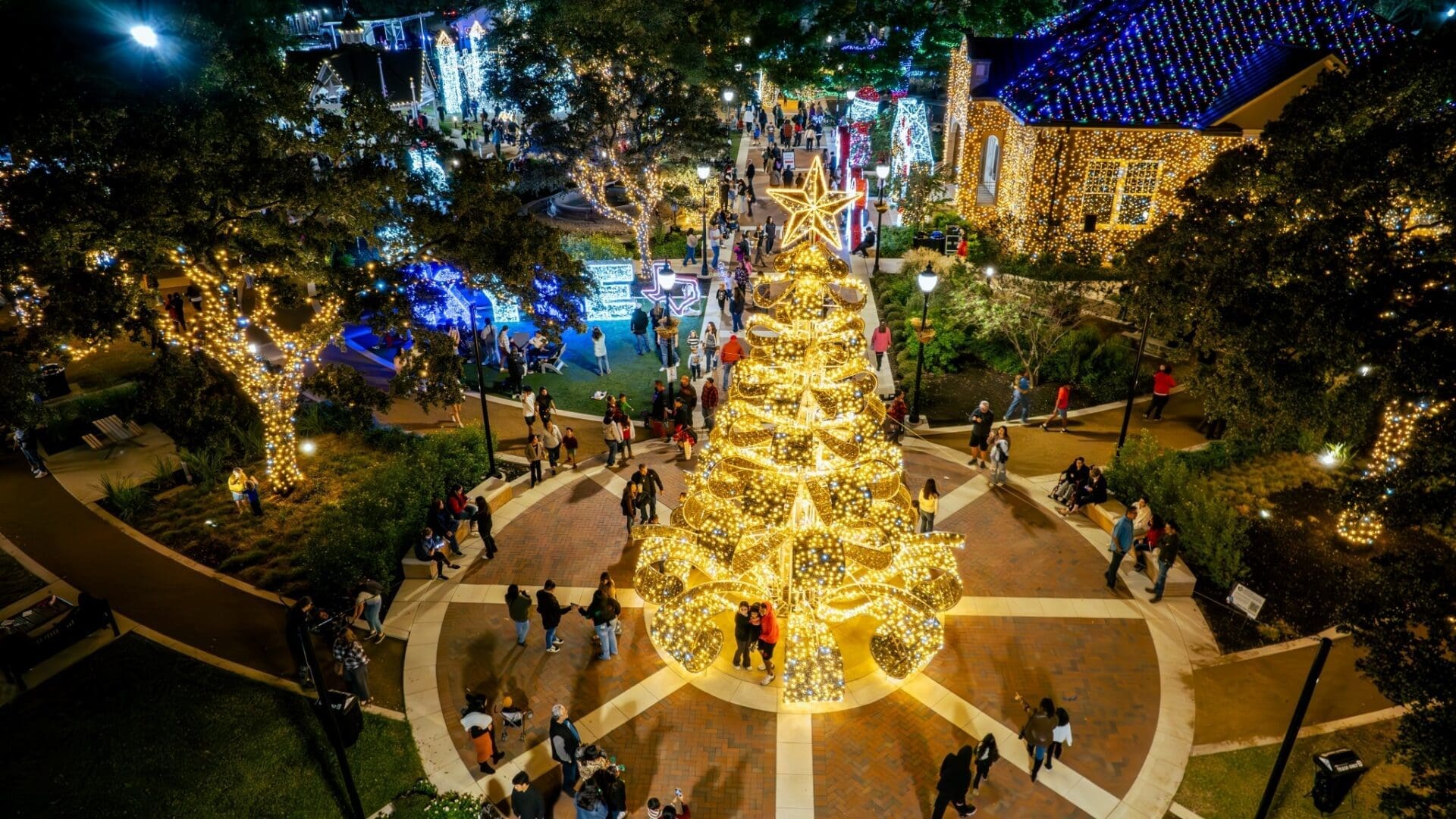 A large Christmas tree adorned with golden lights stands in the center of a lively plaza at night, surrounded by people and festive buildings decorated with bright holiday lights.