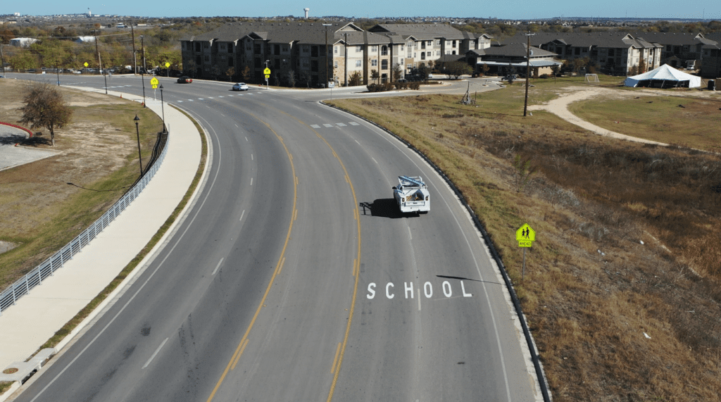 A white truck drives on a wide, empty road near an apartment complex. "SCHOOL" is painted on the street, and a school zone sign is visible on the grassy roadside. The surroundings are open with sparse trees and grass.