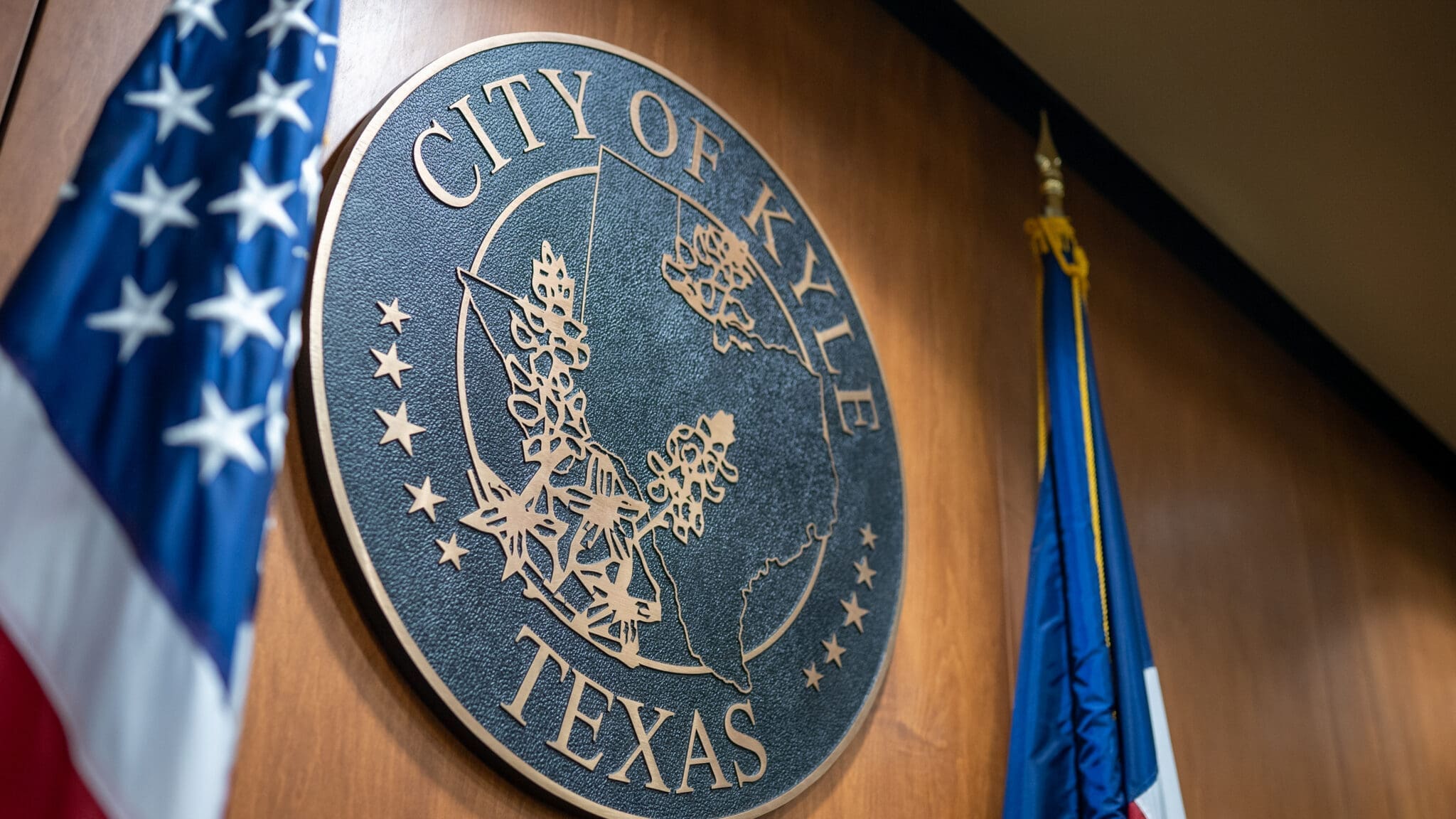 A close-up of the City of Kyle, Texas seal mounted on a wooden wall, with parts of the American and Texas flags visible on either side.