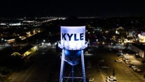 A tall, illuminated water tower labeled “KYLE” stands in a parking lot at night, surrounded by parked cars and city lights in the background.