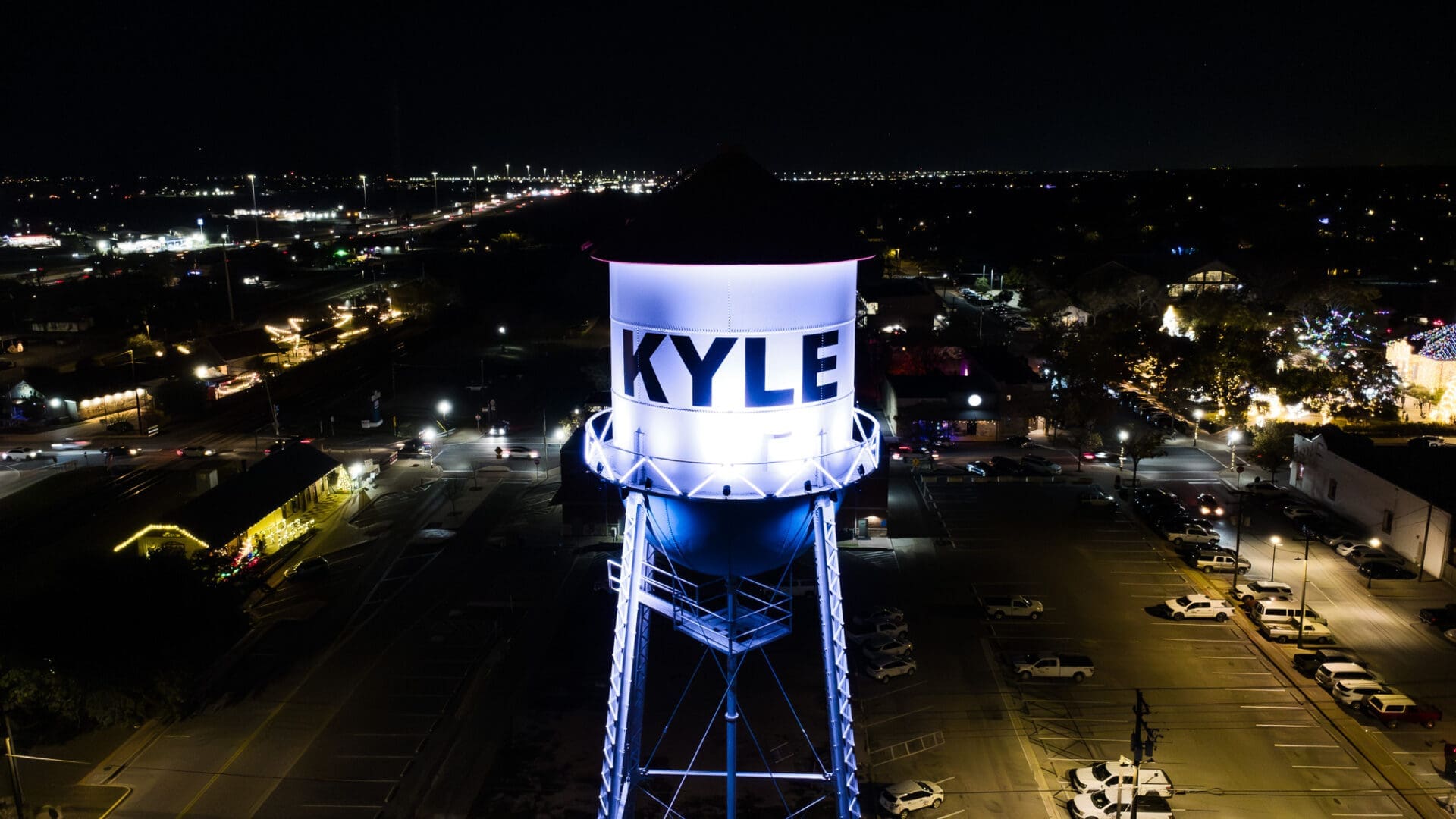 A tall, illuminated water tower labeled “KYLE” stands in a parking lot at night, surrounded by parked cars and city lights in the background.