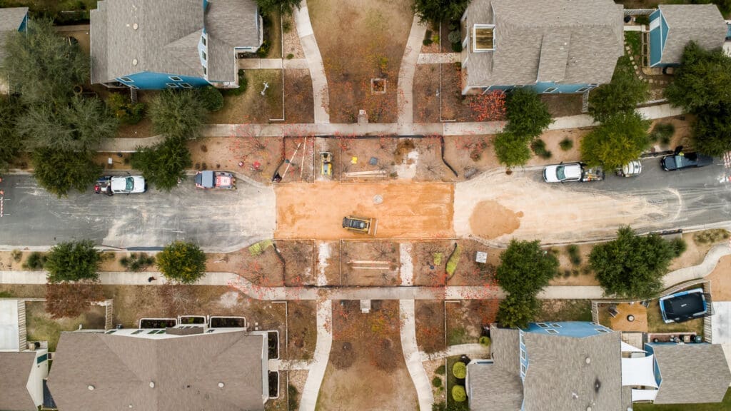 Aerial view of a residential neighborhood with a road under construction; construction vehicles and equipment are present, with orange dirt covering the street and houses lining both sides of the road.