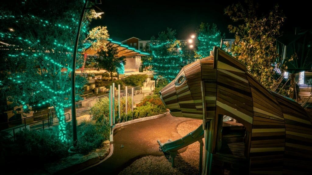 A nighttime park scene with trees wrapped in teal string lights, modern seating areas, and a large wooden animal-shaped play structure in the foreground. Warm lighting highlights pathways and landscaping.