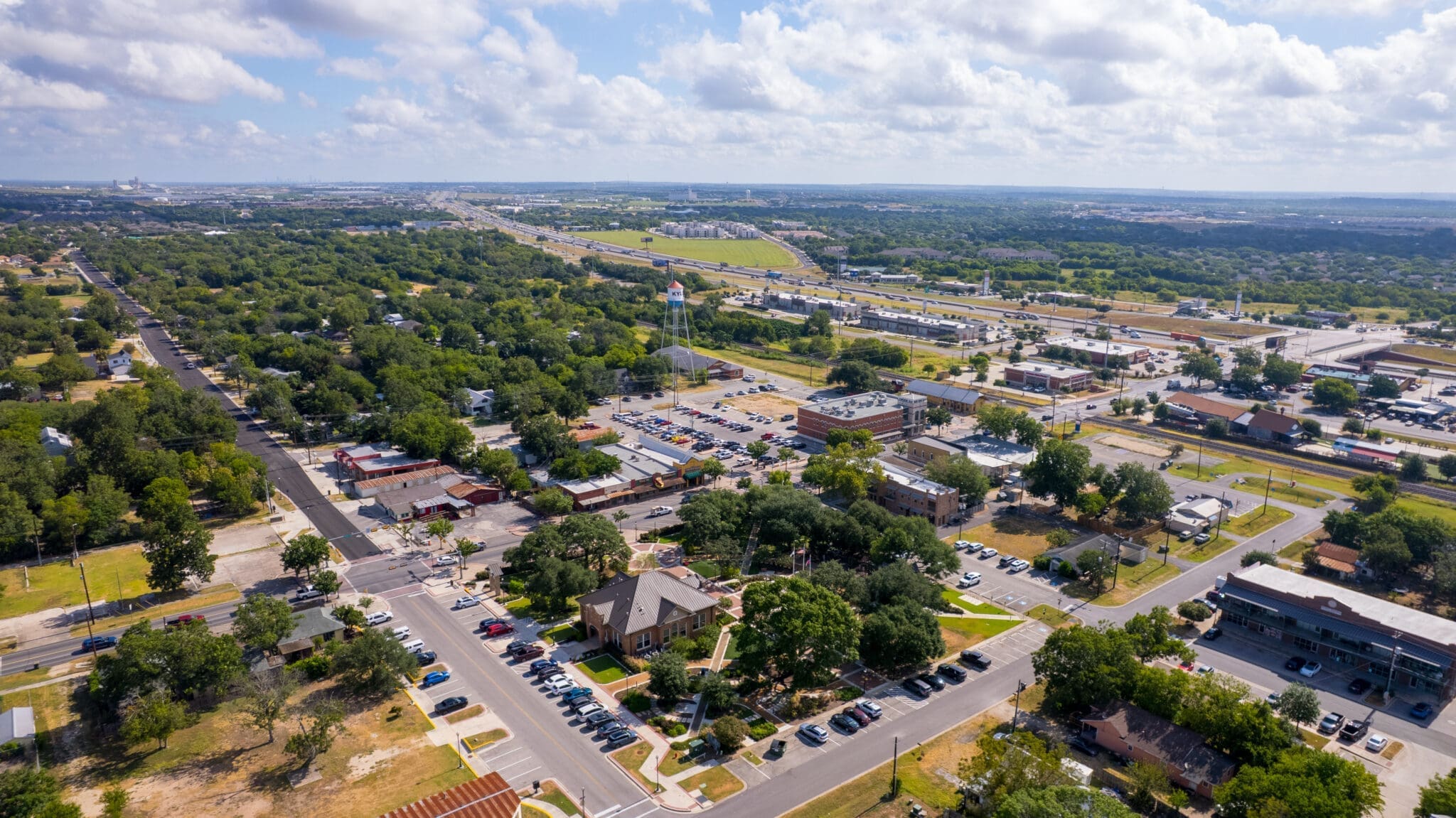 Aerial view of a suburban town with tree-lined streets, residential houses, cars parked, and a highway running through the background under a partly cloudy sky.
