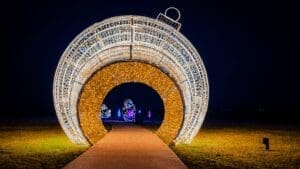 A large, illuminated holiday ornament tunnel made of lights stands over a sidewalk at night, with more colorful light displays and decorations visible in the background.