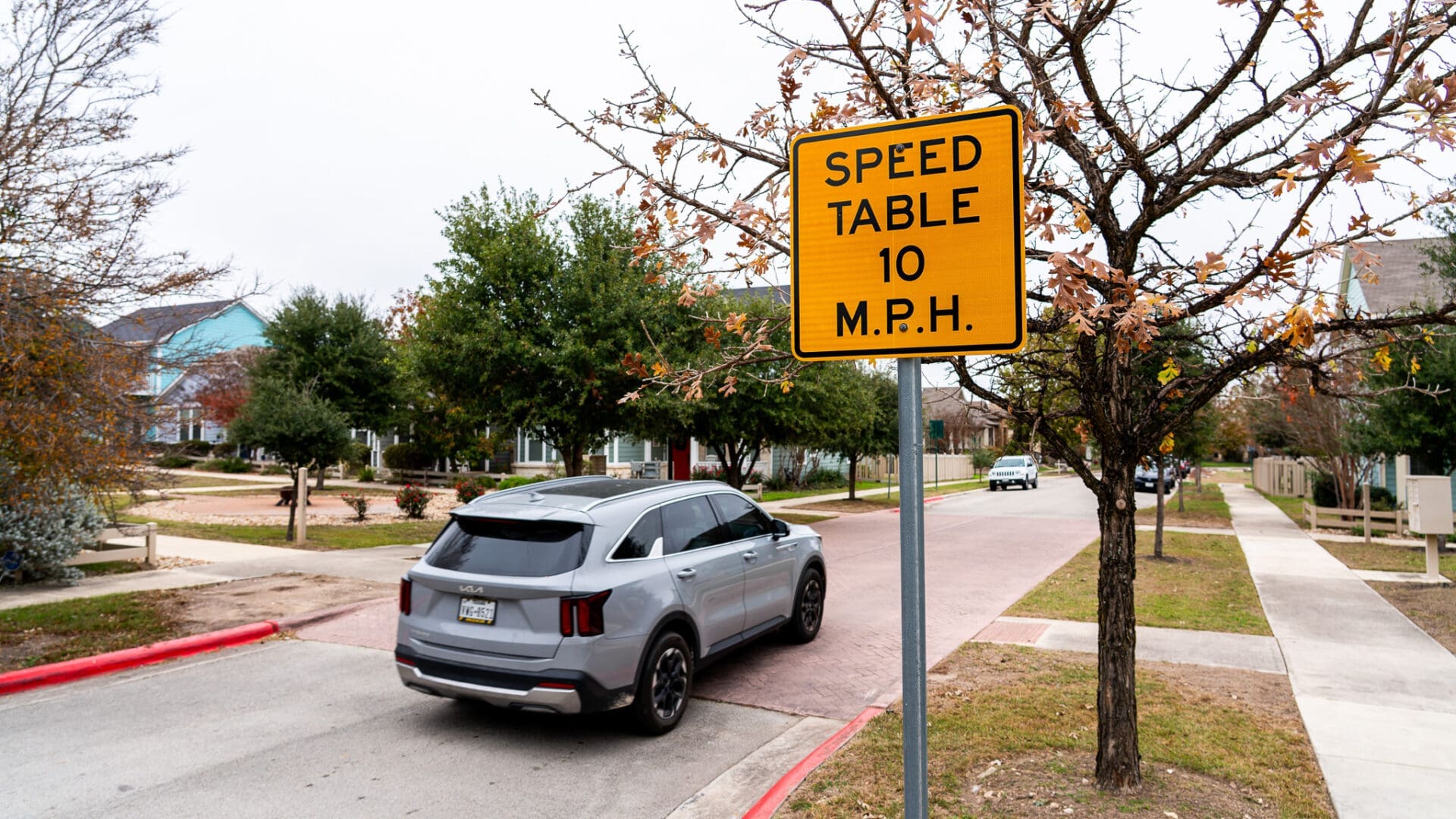 A silver SUV drives past a yellow road sign that reads “Speed Table 10 M.P.H.” on a residential street lined with trees and houses.