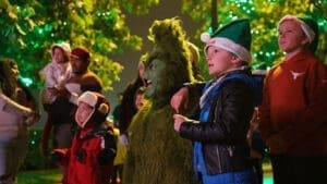 A person dressed as the Grinch stands among children and adults wearing winter clothing and festive hats, surrounded by green holiday lights and trees, enjoying an outdoor event at night.