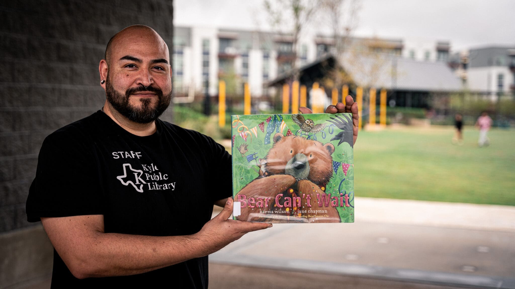A bearded man wearing a black "Staff" t-shirt from Kyle Public Library stands outside and holds up a children's book titled "Bear Can't Wait," smiling at the camera.