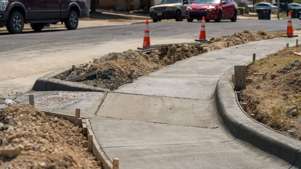 A newly poured concrete sidewalk curves alongside a road under construction, with dirt piles, wooden stakes, and orange traffic cones marking the work zone. Cars are parked on the street nearby.