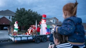 A child on an adult's shoulders watches a festive holiday parade float with Santa, a snowman, a reindeer, and Christmas decorations pass by in a suburban neighborhood at dusk.