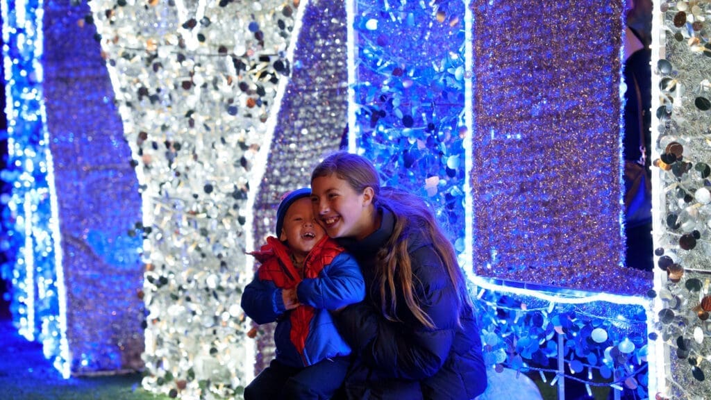 A young woman and a small child in winter jackets smile and pose together in front of large festive blue and white holiday lights and decorations at night.