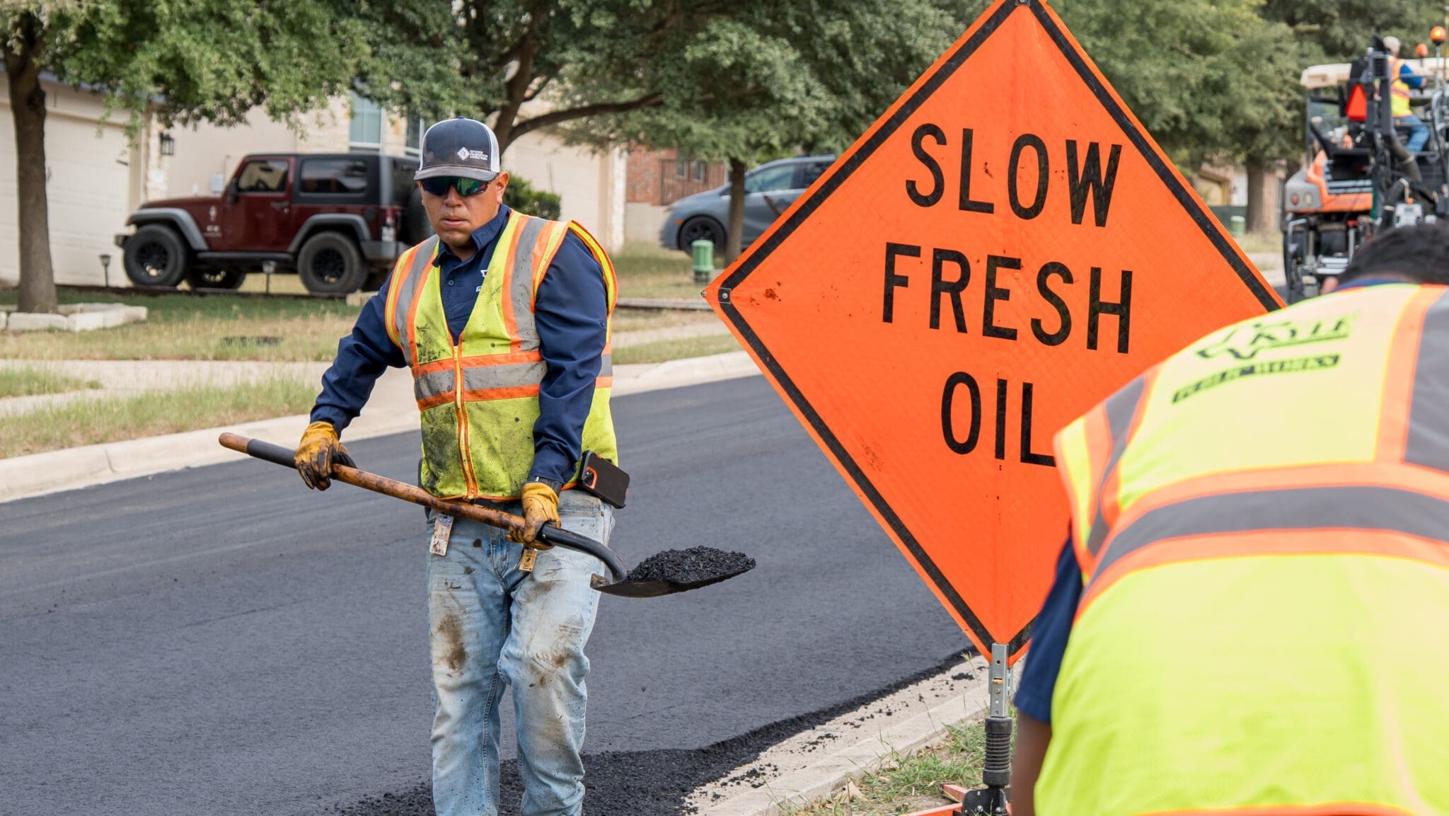 A construction worker in a safety vest and gloves shovels asphalt on a freshly paved road beside an orange sign that reads "SLOW FRESH OIL." Another worker is partially visible in the foreground.
