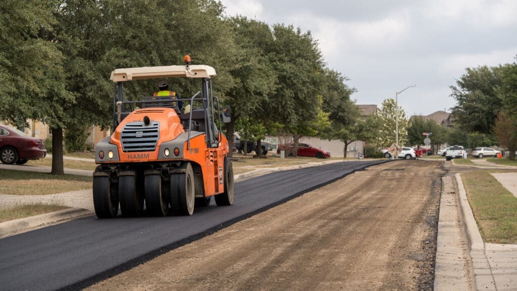An orange road roller compacts fresh asphalt on a residential street, with trees and houses lining both sides and parked cars visible in the background.