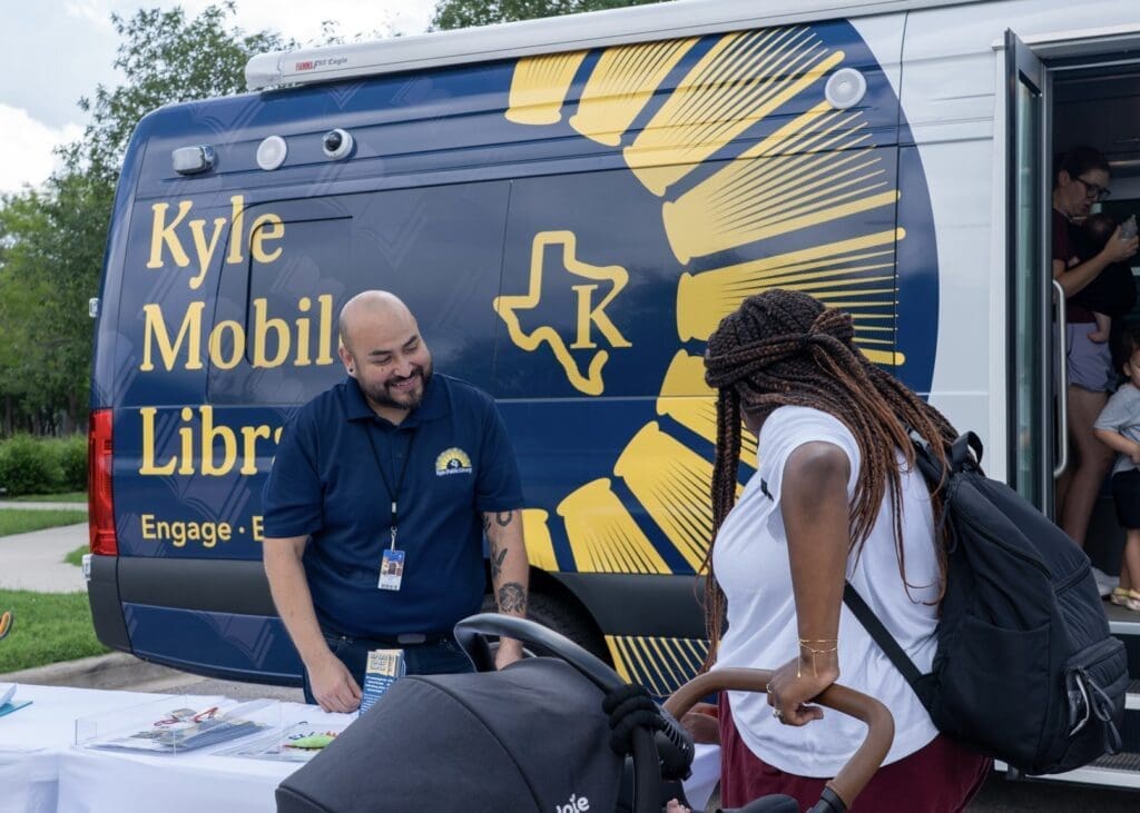 A smiling staff member at the Kyle Mobile Library assists a woman with a stroller at an outdoor event, with the library van and its logo visible in the background.